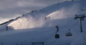 Symbolbild: Mehrere Schneekanonen beschneien einen Skipistenhang; rechts ist ein Skilift zu sehen.